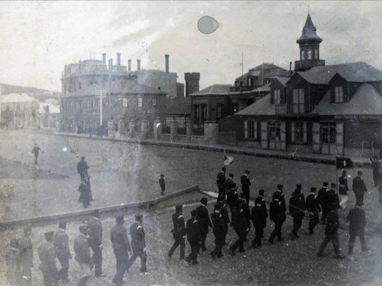 Desfile militar en la esquina de la plaza de Punta Arenas, con construcciones de familias Braun y Menéndez